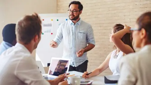 Schulungen im Team – fünf Personen in einem Besprechungsraum, eine Person steht und spricht, andere hören zu. Whiteboard im Hintergrund