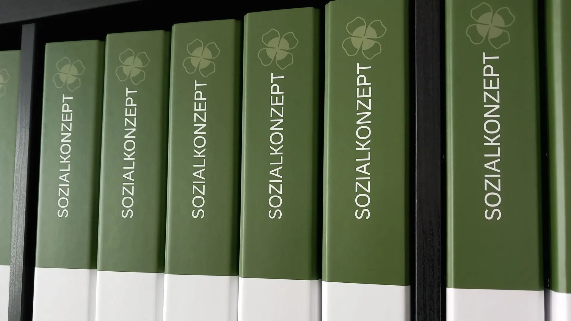 Grün-weiße Ordner mit der Aufschrift ‚Sozialkonzept‘ und Kleeblatt-Symbol im Regal – symbolisieren strukturierte Erstellung von Sozialkonzepten