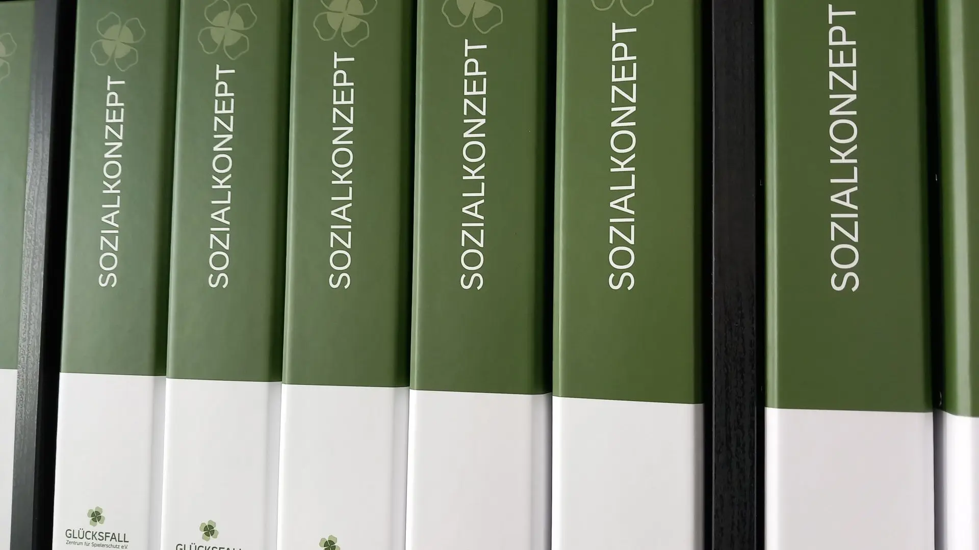 Sozialkonzept – symbolisch dargestellt durch beschriftete Ordner mit Glücksfall-Logo Grüne und weiße Ordner mit der Aufschrift ‚Sozialkonzept‘ und Kleeblatt-Symbol im Regal – symbolisieren strukturierte Erstellung von Sozialkonzepten.