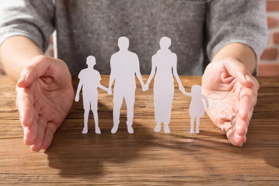 Tipps für Angehörige – Symbolbild mit Familie und schützenden Händen Symbolbild für Unterstützung – Papiersilhouetten einer Familie auf Holzuntergrund, umrahmt von schützenden Händen.